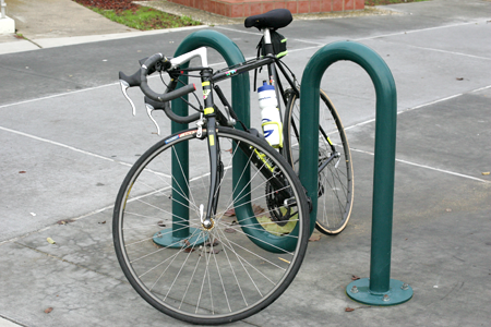 A black road bicycle locked to a green inverted-U style bike rack on a concrete sidewalk